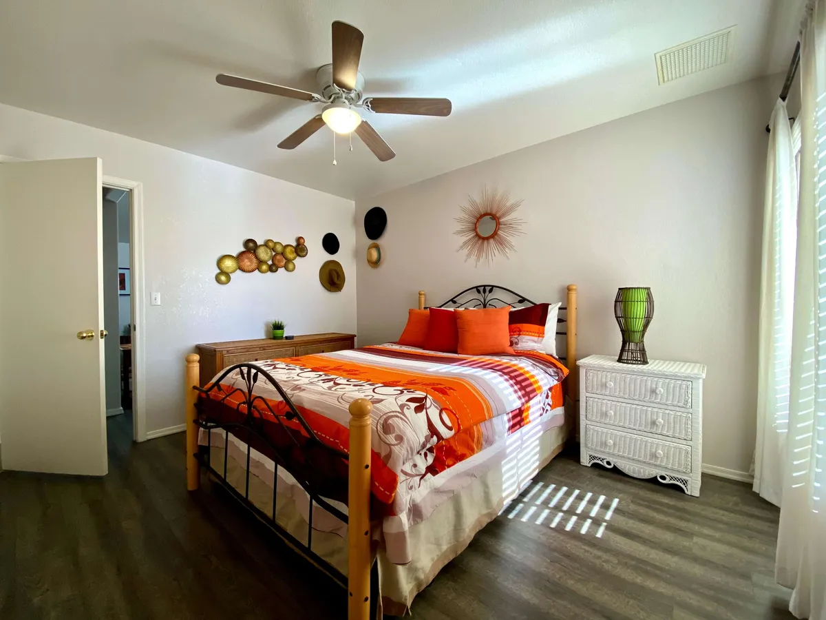 The image shows a bright and inviting bedroom with vibrant bedding, decorative wall art, and a crisp white wicker dresser. Sunlight pours through the window, creating a cheerful, welcoming atmosphere that suggests both relaxation and togetherness. The room balances fresh, modern touches with cozy comfort—perfect for unwinding or connecting with loved ones.