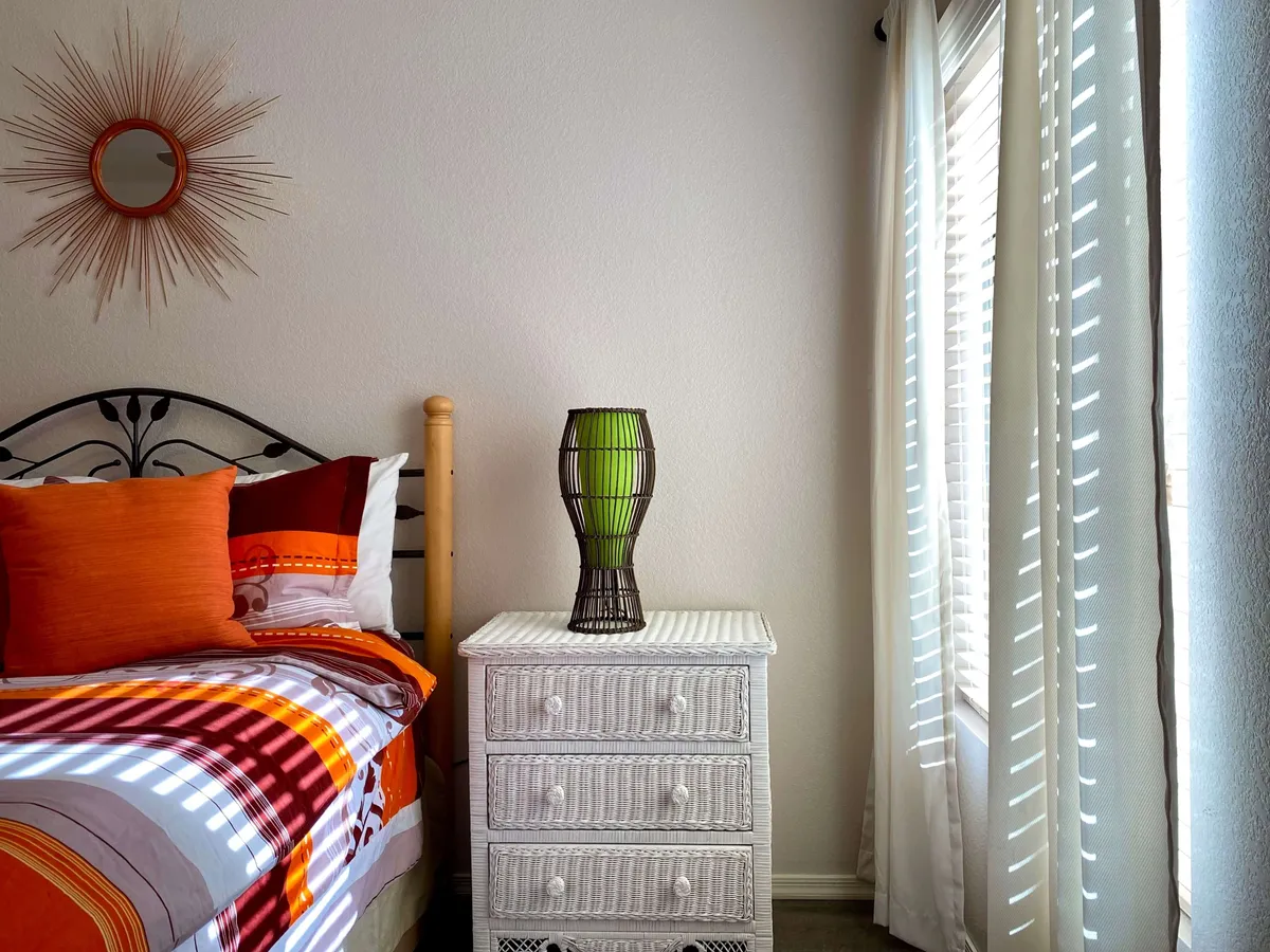 A cheerful guest bedroom featuring a bed adorned with colorful bedding, a green lamp on the nightstand, and a decorative sunburst mirror on the wall. The window fills the space with soft morning light, creating a cozy and inviting ambiance.