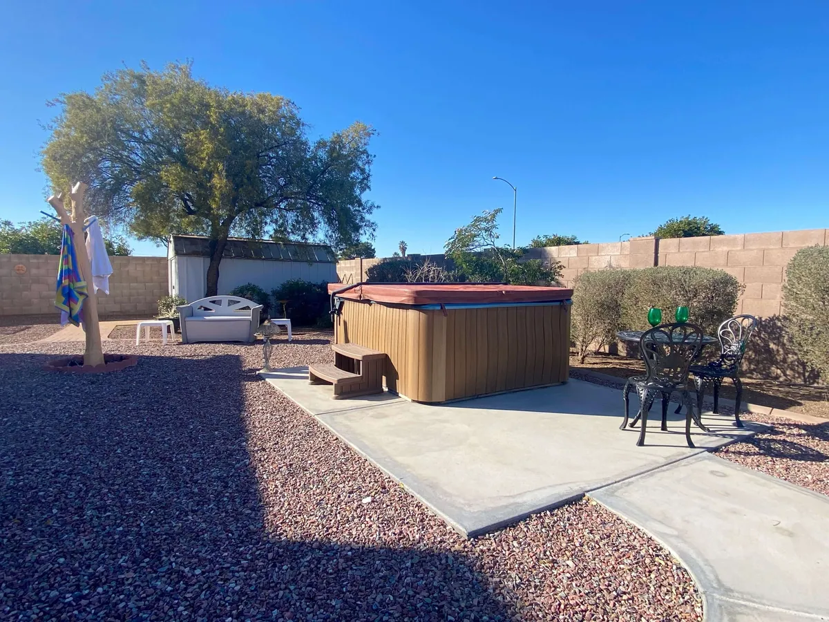 A serene backyard scene featuring a spacious hot tub surrounded by seating, a towel rack, a large tree, and lush landscaping. The area is designed for relaxation with space for drinks and socializing, suggesting both comfort and privacy.