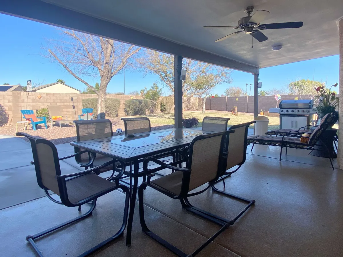 This image shows a backyard setup featuring a dining table with chairs, a grill, lounge chairs, and a lush backyard view. The scene is bathed in sunlight, suggesting opportunities for outdoor dining, barbecuing, and relaxed socializing in a private outdoor space.