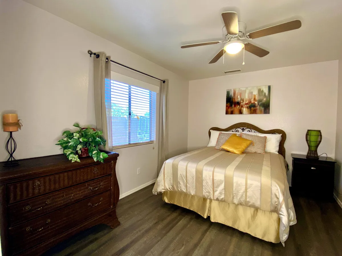 A comfortable bedroom with a cozy bed, dresser, window allowing natural light, a ceiling fan for added comfort, and tasteful artwork. The soft, inviting styling makes this room ideal for relaxation and restful sleep.