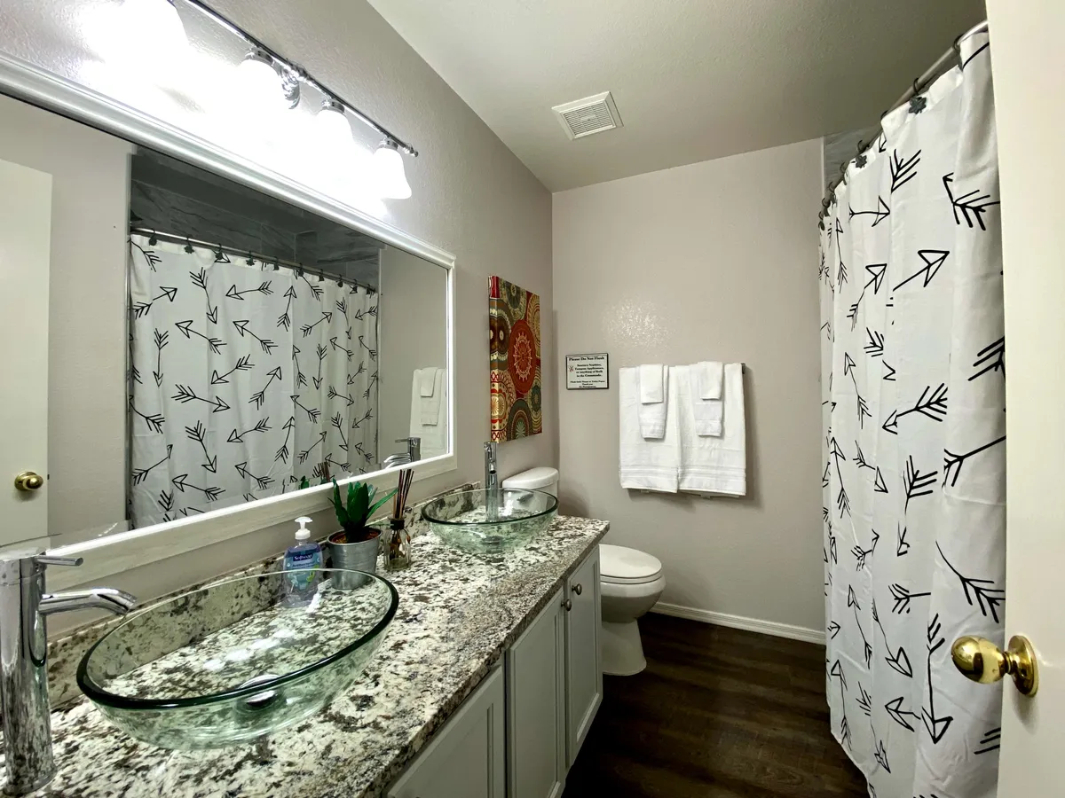 A serene bathroom with dual glass vessel sinks on a granite countertop, stylish decorative wall art, and a shower with a patterned curtain. The bright, clean design exudes a spa-like feel ideal for morning and evening routines.