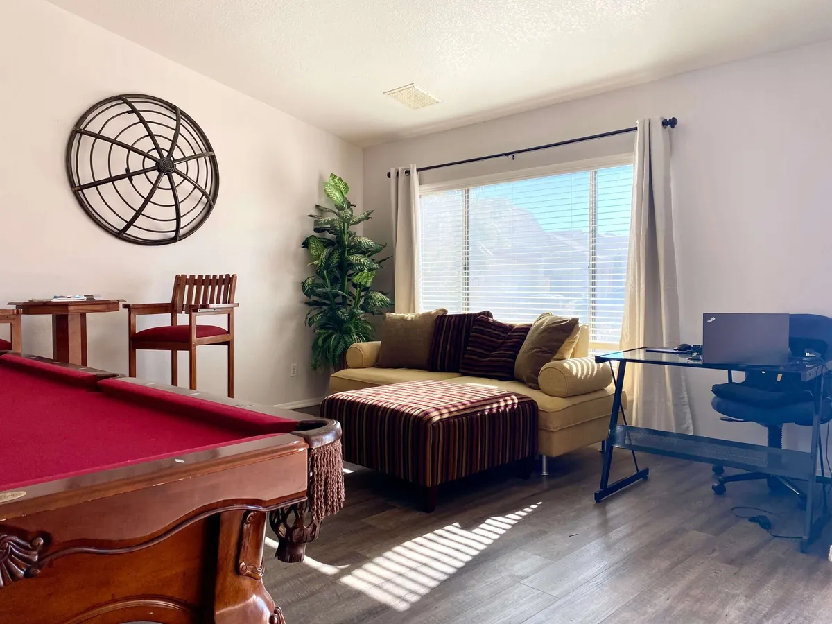 A versatile interior space featuring a pool table, a comfortable seating area, a desk with a computer, and a window streaming in natural light. The setup combines relaxation, socialization, and a dedicated workspace, suggesting the dual purpose of work and play.