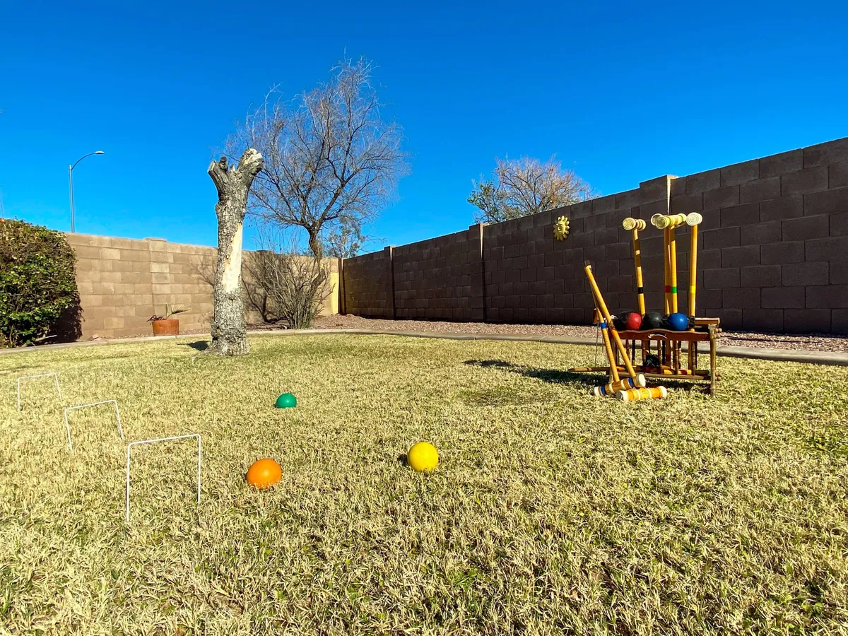 A cheerful backyard scene with a croquet set, colorful balls, a large shade tree, and a fenced yard. The space is clearly designed for active outdoor play and family-friendly leisure, inviting both children and adults to participate in classic yard games.