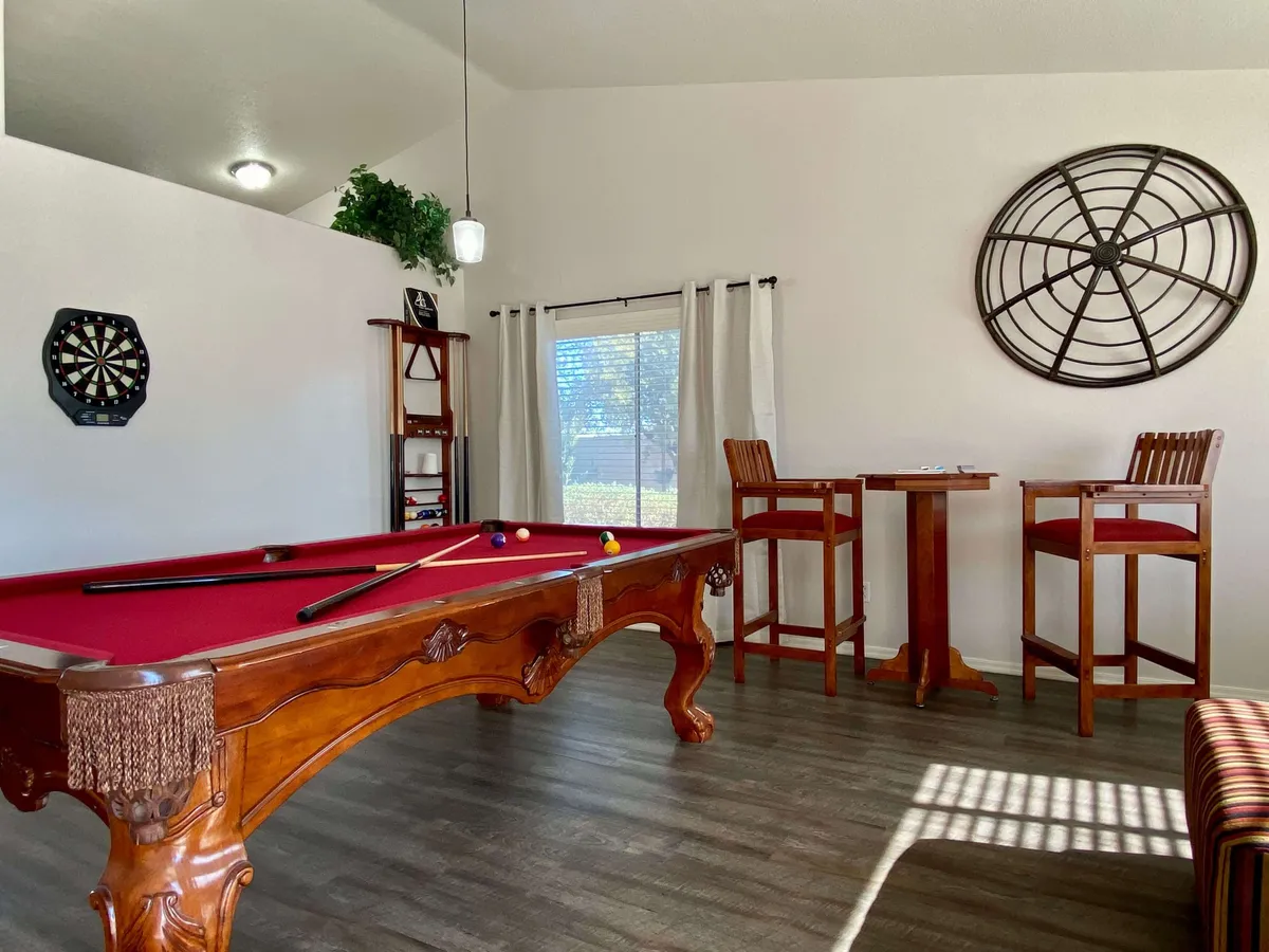 A vibrant game room spotlighting a full-size pool table, a dartboard mounted on the wall, high-top tables for drinks or games, and a decorative wall clock. A large window brings in natural light, while the casual furnishings invite guests to enjoy friendly competition or socialize in a lively atmosphere.